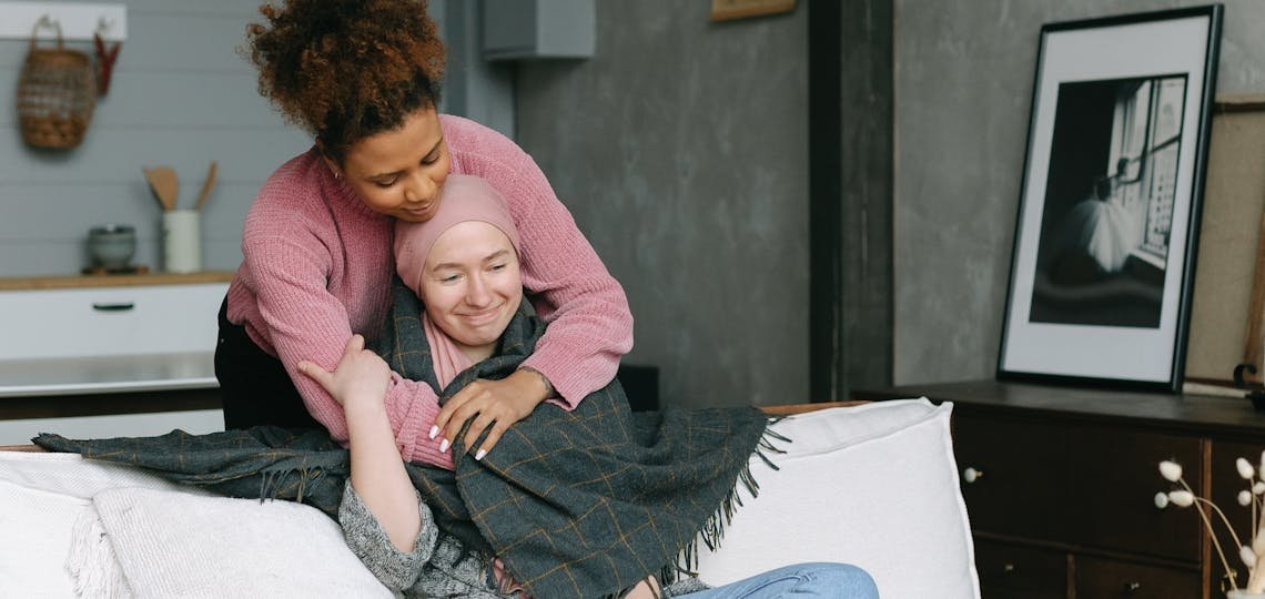 Two women share a caring embrace in a cozy, modern living room, embodying support for a terminally ill cancer sufferer