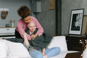 Two women share a caring embrace in a cozy, modern living room, embodying support for a terminally ill cancer sufferer