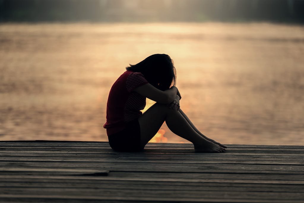 A woman sitting alone on a wooden dock by the lake, showing solitude and grief.