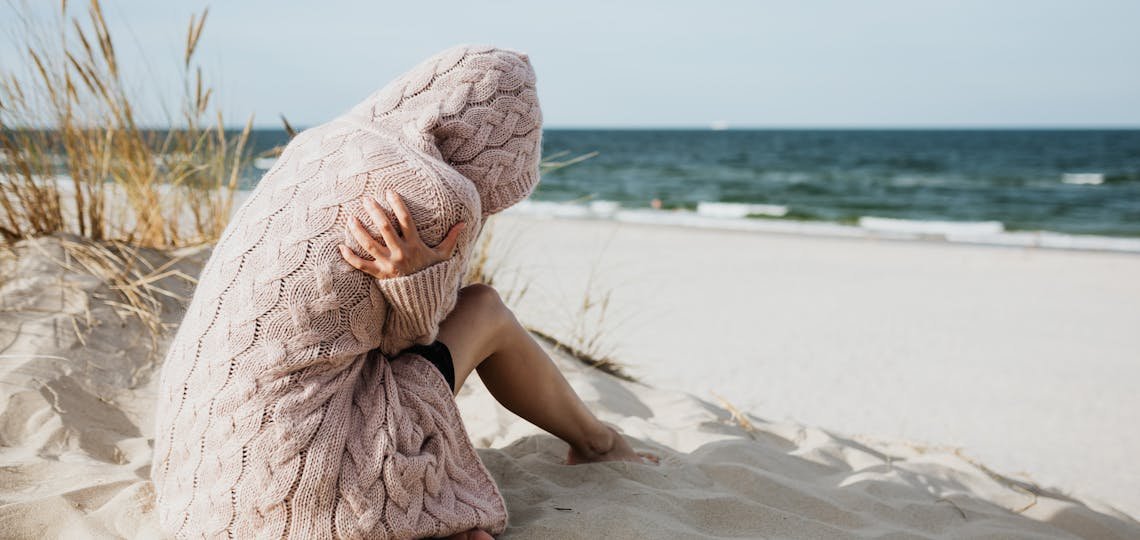 Solitary woman in pink knitted sweater on a tranquil beach setting with her arms wrapped around her as she comes to terms with guilt and grief.