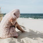Solitary woman in pink knitted sweater on a tranquil beach setting with her arms wrapped around her as she comes to terms with guilt and grief.