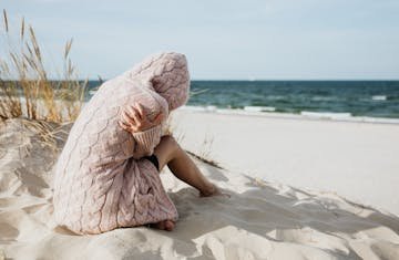 Solitary woman in pink knitted sweater on a tranquil beach setting with her arms wrapped around her as she comes to terms with guilt and grief.