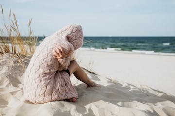 Solitary woman in pink knitted sweater on a tranquil beach setting with her arms wrapped around her as she comes to terms with guilt and grief.