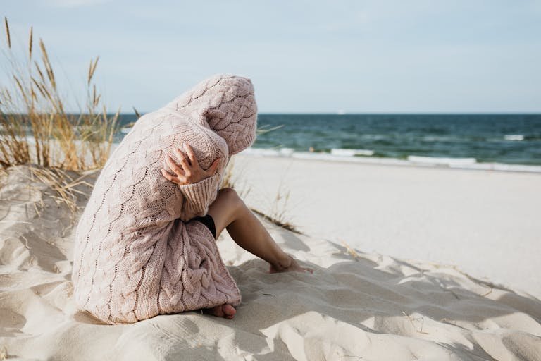 Solitary woman in pink knitted sweater on a tranquil beach setting with her arms wrapped around her as she comes to terms with guilt and grief.