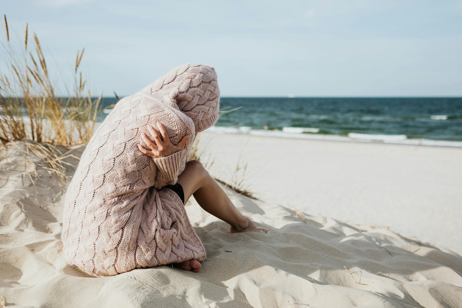 Solitary woman in pink knitted sweater on a tranquil beach setting with her arms wrapped around her as she comes to terms with guilt and grief.