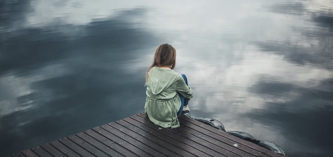 A solitary woman in a green hoodie sits on a wooden dock, reflecting silently by a placid lake.
