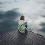 A solitary woman in a green hoodie sits on a wooden dock, reflecting silently by a placid lake.