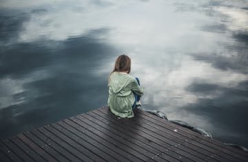 A solitary woman in a green hoodie sits on a wooden dock, reflecting silently by a placid lake.