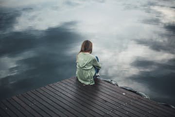 A solitary woman in a green hoodie sits on a wooden dock, reflecting silently by a placid lake.